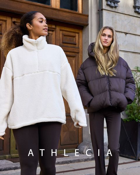 Deux femmes en vêtements de sport Athlecia bleus posent sur un terrain rocailleux. L'une porte un sweat-shirt et un short, l'autre un crop-top blanc et un pantalon bleu. Au-dessus d'elles, on peut voir le nom de la marque « Athlecia ».