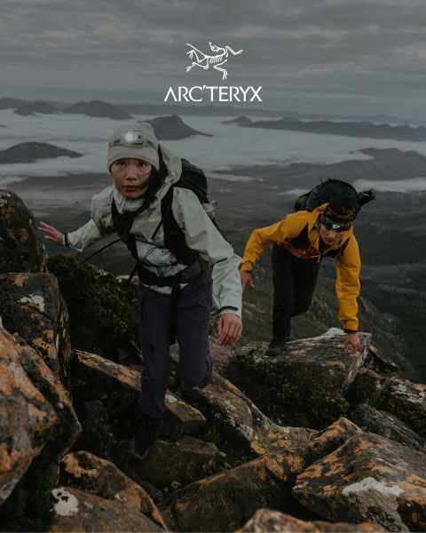 Deux personnes randonnent sur un sentier de montagne rocailleux. Le ciel est couvert, une chaîne de montagnes brumeuse est visible en arrière-plan. Les deux randonneurs portent des équipements de plein air. Un petit étang se trouve à droite. Le logo Arc'teryx est visible en haut.