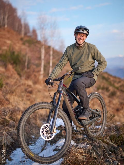 Une personne avec un casque et une veste verte sourit en posant avec un VTT sur un chemin rocheux et enneigé, avec des arbres en arrière-plan.