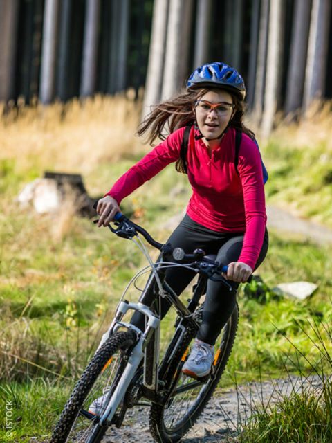 Une femme en chemise rouge et avec un casque fait du VTT sur un chemin forestier ensoleillé.