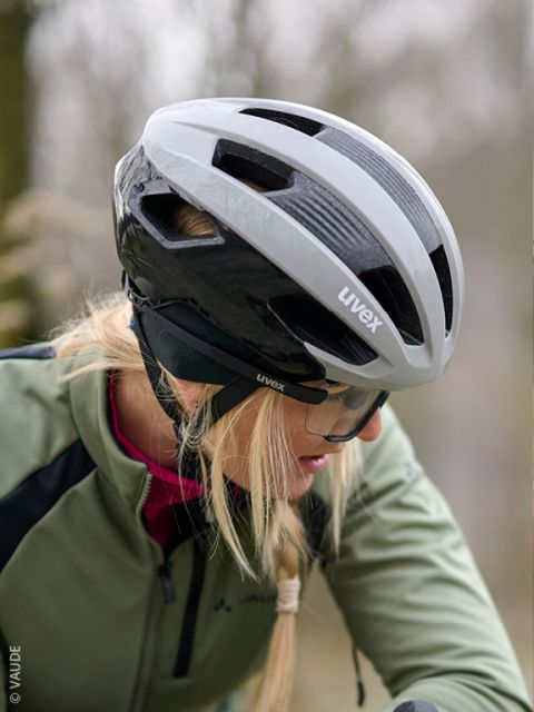 Un homme en veste verte et gants examine un casque de vélo noir et est assis devant un mur en bois. Il porte un bandeau noir avec une inscription blanche.