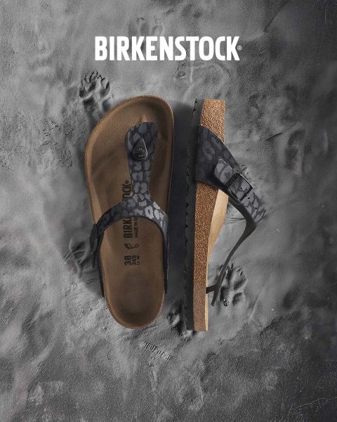 Une personne portant des sandales Birkenstock beiges se tient à côté d'un jeu d'eau dans un paysage de jardin luxuriant. La zone est entourée de verdure et le mot « BIRKENSTOCK » est clairement visible au-dessus de l'image.
