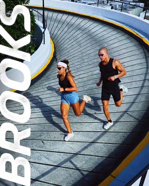 Un groupe de cinq personnes traverse énergiquement un passage piéton. Ils portent des vêtements de sport colorés, notamment des shorts et des débardeurs. La scène est animée, et le mot « BROOKS » est bien visible au-dessus d'eux.