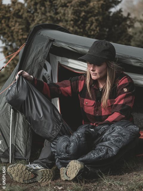 Une femme en chemise à carreaux rouge est assise devant une tente, enfilant un pantalon de pluie et des chaussures de randonnée, se préparant pour des activités de plein air.