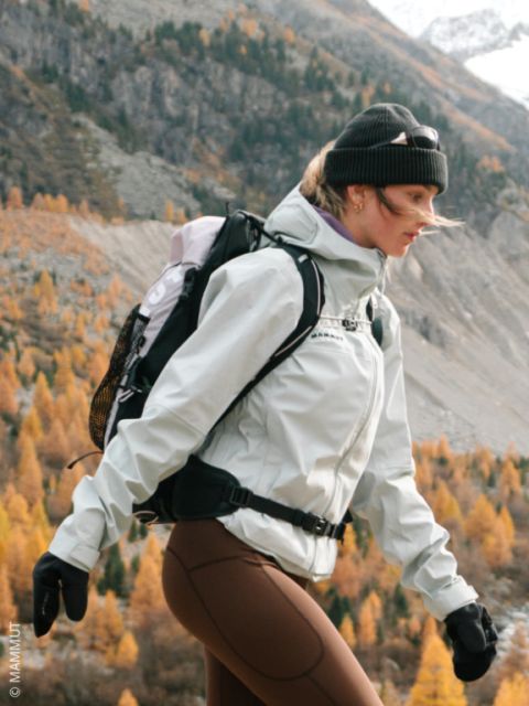 Une femme fait de la randonnée en automne dans la nature, vêtue d'une veste, d'un bonnet, de gants et d'un sac à dos. À l'arrière-plan, on peut voir des montagnes et des orangers.