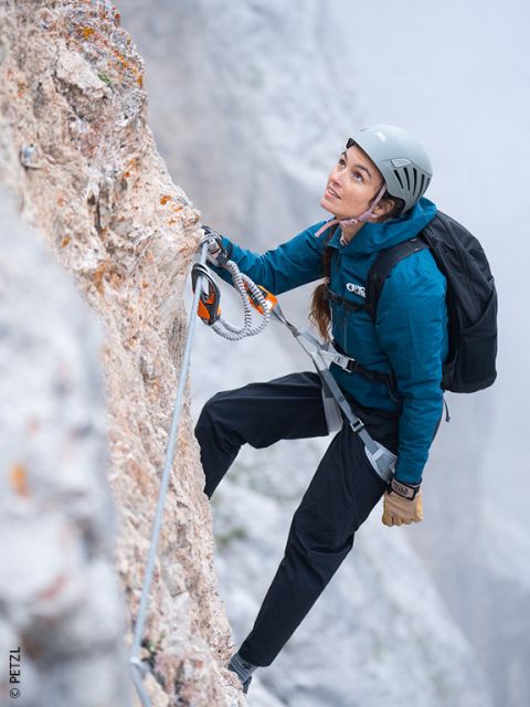 Une femme en équipement d'escalade grimpe une falaise rocheuse, sécurisée par une corde de sécurité, regardant vers le haut avec concentration. Il y a du brouillard en arrière-plan.