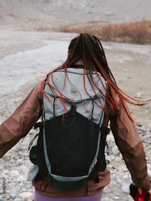 Une personne aux cheveux tressés et portant un grand sac à dos marche sur un terrain rocheux près d'une rivière, le dos tourné vers la caméra.