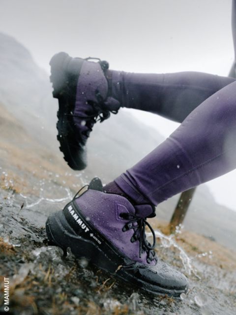 Gros plan sur les jambes d'une personne portant un pantalon violet et des chaussures de randonnée noires, pataugeant dans une flaque d'eau sur un sentier de randonnée rocheux et mouillé par la pluie.