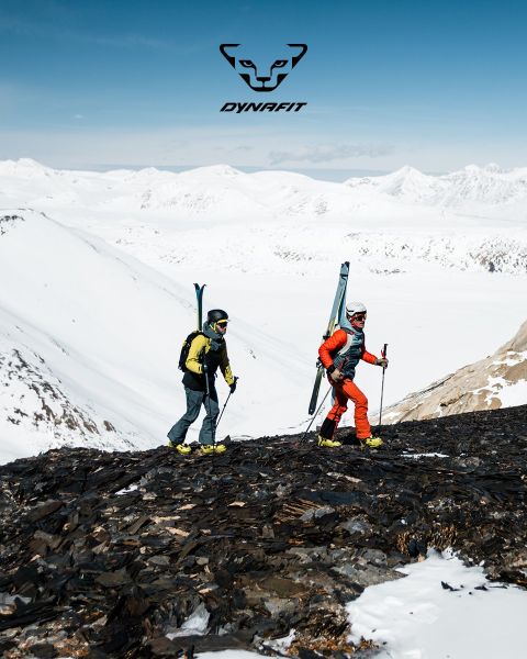 Deux personnes courent sur un sentier de montagne avec une ville et une rivière en arrière-plan. L'une porte un t-shirt noir et un short, l'autre un débardeur bleu et un short. Le logo Dynafit se trouve en haut de l'image.