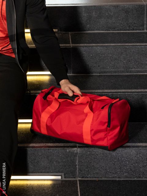 Une personne en jogging beige clair, baskets noires et haut noir tient un sac de sport noir brillant. Elle se tient sur des planches de bois foncé à côté d'un vieux projecteur et de valises empilées.