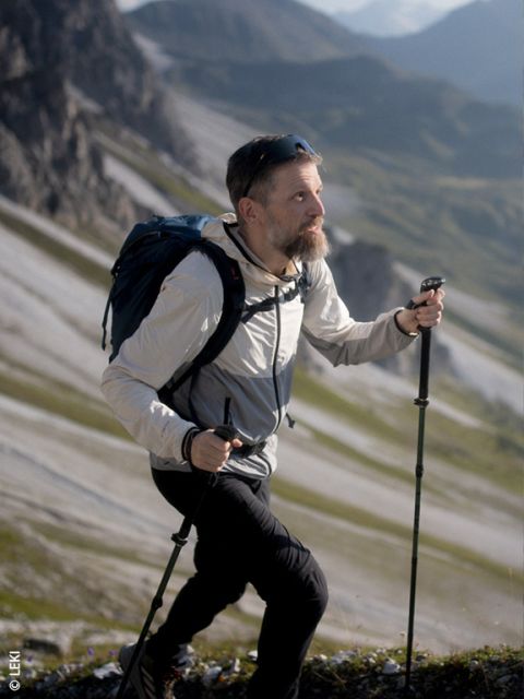Un homme barbu marche en montée sur un sentier de montagne rocheux, utilisant des bâtons de randonnée et portant un sac à dos ; des montagnes sont visibles en arrière-plan.