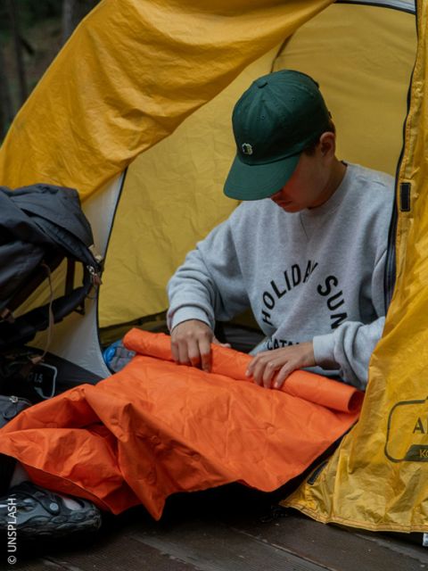Une personne portant un bonnet vert est assise dans une tente jaune et plie un tapis de sol orange, alors qu'elle campe en plein air.
