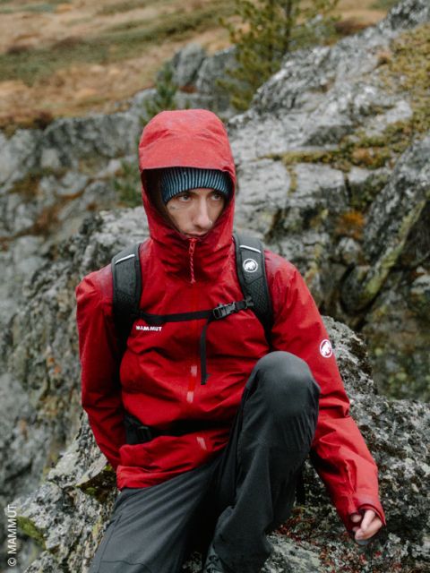 Une personne vêtue d'un sweat à capuche rouge et d'un sac à dos est assise dehors sur des rochers, avec des arbres et un terrain rocheux en arrière-plan.