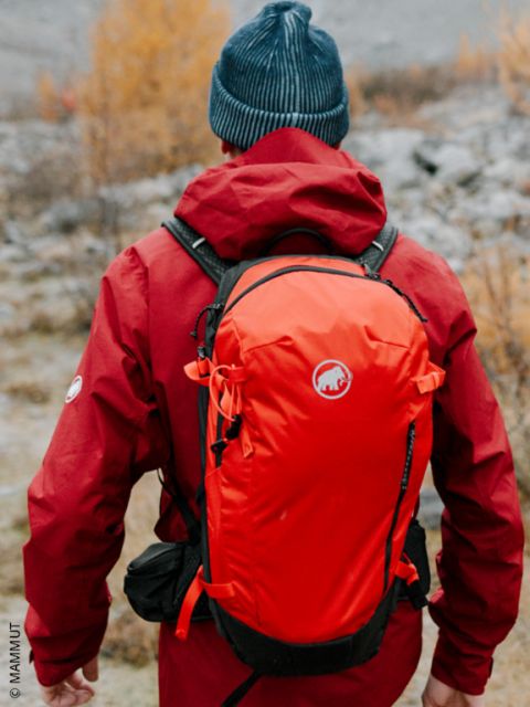 Une personne en veste rouge et bonnet bleu, faisant de la randonnée dans la nature avec un sac à dos orange, vue de dos.