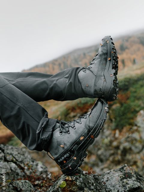 Une personne en bottes de randonnée noires, couvertes de terre et de feuilles, repose ses pieds sur des rochers en plein air, avec un paysage montagneux flou en arrière-plan.