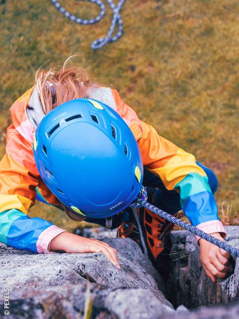 Une personne avec un casque bleu et une veste colorée escalade une paroi rocheuse, sécurisée par une corde. Vue d'en haut, un sol herbeux est visible en dessous.