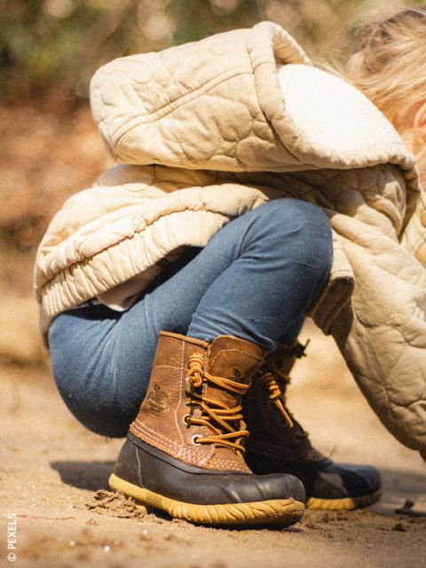 Un petit enfant en manteau beige et bottes marron est accroupi dehors sur un sol sablonneux, partiellement tourné dos à la caméra.