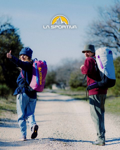 Quatre personnes en équipement de plein air, avec des tapis de bloc sur le dos, montent un sentier de montagne herbeux. Au-dessus d'eux, le logo de La Sportiva est visible dans le ciel. À l'arrière-plan, on aperçoit des sommets montagneux escarpés.