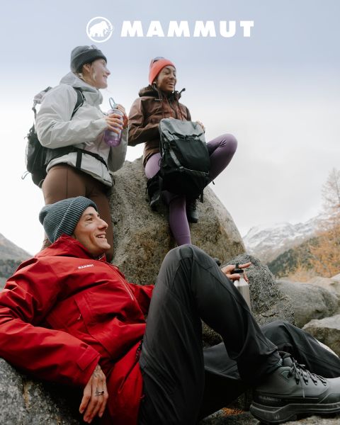 Deux randonneurs en équipement de plein air traversent un terrain rocailleux sous un ciel nuageux, avec une grande montagne en arrière-plan. Ils portent tous les deux des vêtements Mammut, et le logo de la marque est visible en haut de l'image.