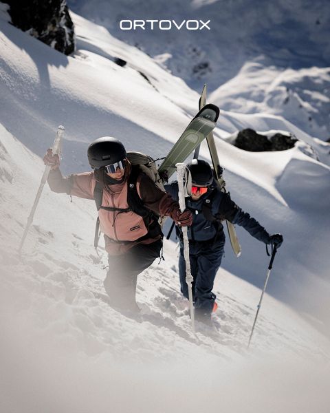 Trois personnes en équipement de plein air sont assises, souriantes et discutant sur un terrain rocheux dans une région montagneuse. Le soleil se couche derrière les sommets. Un sac à dos et une thermos sont posés à proximité. Le logo d'Ortovox se trouve en haut à droite.