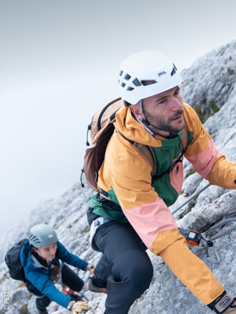 Un alpiniste avec un casque, des lunettes de soleil et du matériel d'escalade gravit une pente rocheuse avec des sommets enneigés en arrière-plan sous un ciel clair.