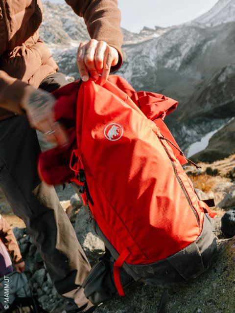 Une personne en vêtements d'hiver et gants à motifs place une pelle à neige dans un sac à dos rose sur un sol enneigé, avec des arbres enneigés en arrière-plan.