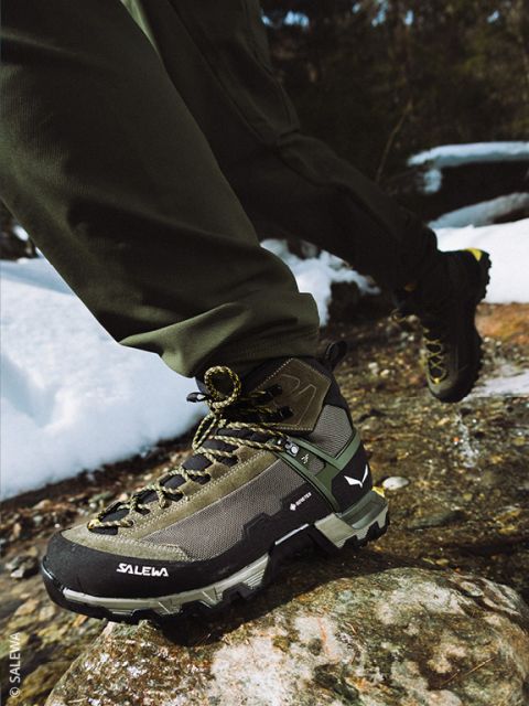 Une personne en tenue de plein air serre les lacets d'une chaussure de randonnée noire, debout sur une surface rocheuse, avec une grande formation rocheuse et un ciel bleu en arrière-plan.
