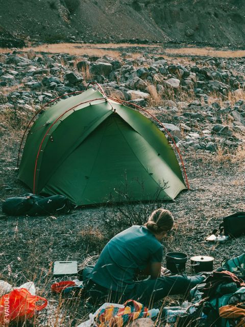 Une personne en veste rouge et bonnet prépare de la nourriture à l'extérieur, accroupie dans l'herbe, près de matériel de camping, vue à travers la porte ouverte d'une tente avec des montagnes et des arbres en arrière-plan.