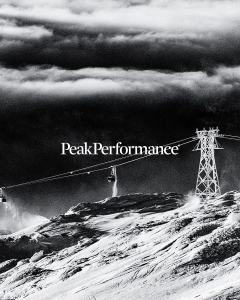 Une personne saute entre de grands rochers dans une région montagneuse sous un ciel bleu éclatant. Elle porte des vêtements de sport, notamment une veste et un short. En haut, on peut lire l'inscription « Peak Performance ».