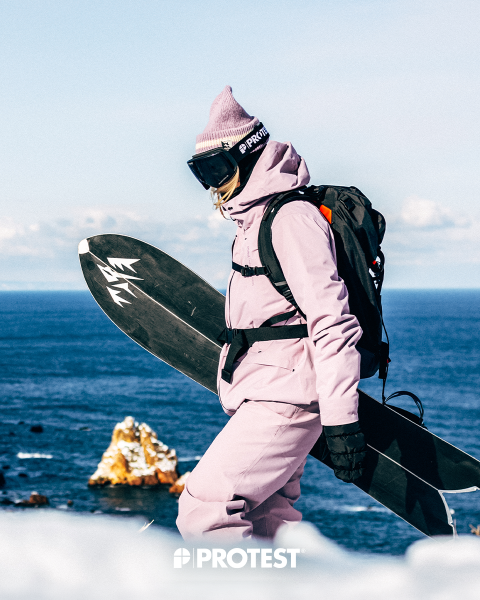 Une personne en combinaison de ski rose et bonnet, tenant un snowboard, se tient au bord de la mer, avec des formations rocheuses en arrière-plan et le mot « PROTEST » en bas.