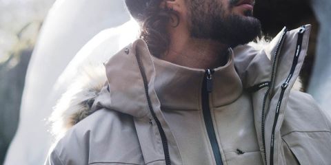 Un homme en veste marron, pantalon noir et bonnet gris se promène dans une forêt dense. Il porte des gants noirs et un sac à dos. La forêt a de grands arbres et des feuilles tombées. Logo « Rukka » en haut.