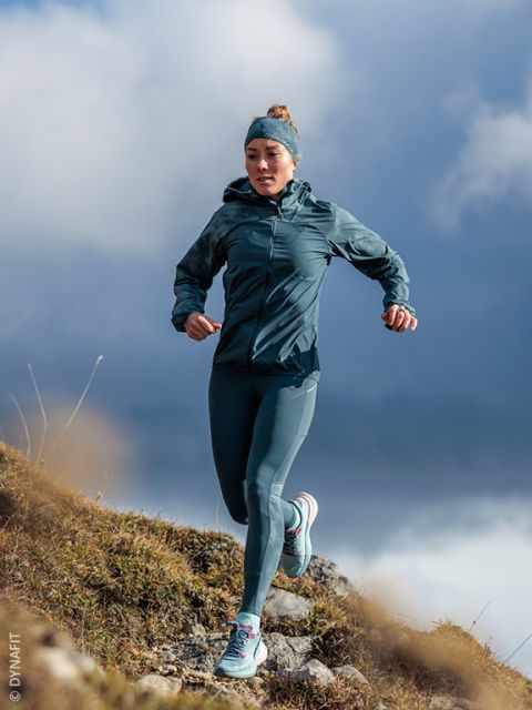 Une femme en tenue de sport turquoise court en descente sur un sentier rocheux, avec un ciel nuageux en arrière-plan.