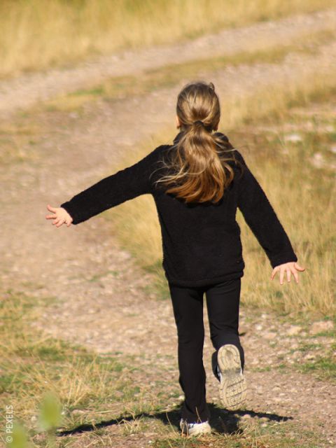 Une fille aux cheveux longs vêtue d'une tenue noire court joyeusement les bras tendus le long d'un chemin de terre bordé d'herbe.