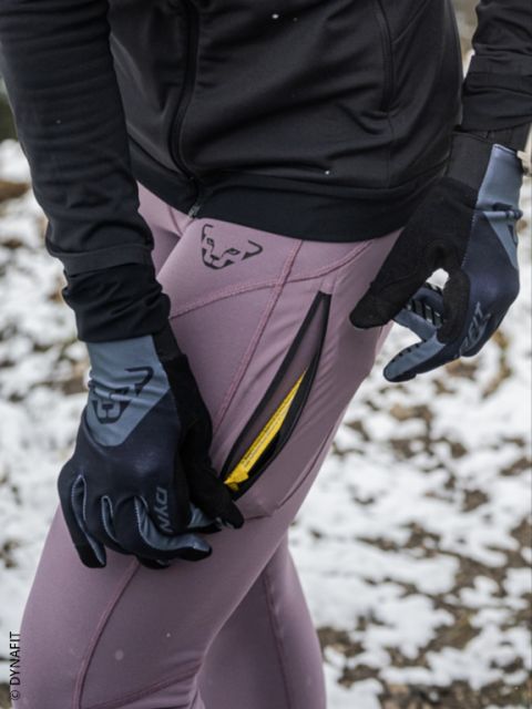 Deux personnes courant de nuit en tenue de sport, avec des gouttes de pluie visibles dans l'air et un trottoir mouillé sous leurs pieds. Les deux coureurs portent des vestes et des chaussures de course et passent rapidement devant un portail métallique.