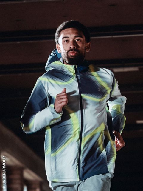 Une personne portant un sweat à capuche et des chaussures de course fait du jogging sur un chemin enneigé par une journée froide et claire, avec la ligne d'horizon de la ville en arrière-plan.