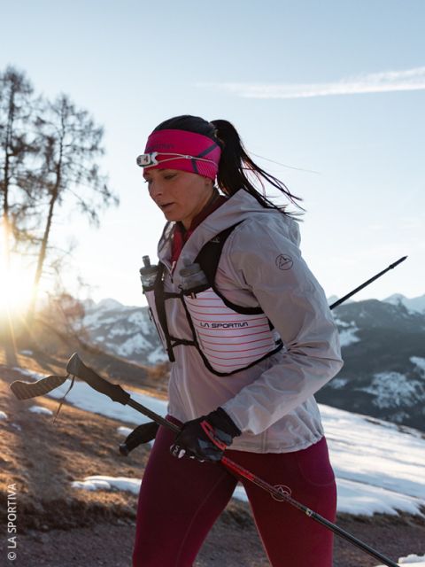 Une femme portant une veste et des gants rose-gris court dans un paysage montagneux enneigé, transportant deux bouteilles d'eau dans les poches avant de son gilet de course.