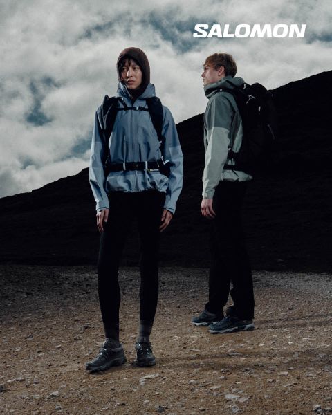 Gros plan d'une personne portant des chaussures Salomon et des chaussettes Salomon avec un dégradé de couleurs bleues. Elle se tient dans une prairie avec un paysage de montagne pittoresque en arrière-plan. Un randonneur en vêtements d'extérieur se tient plus loin, également dans la prairie.