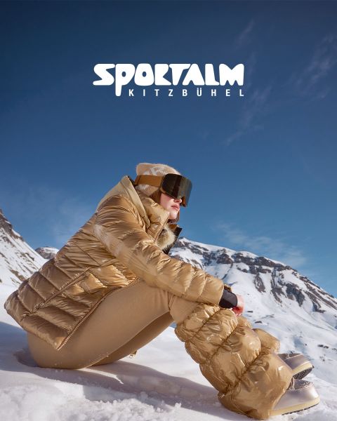 Une personne portant une doudoune métallique et un pantalon noir est allongée sur un terrain enneigé avec des montagnes en arrière-plan sous un ciel nuageux. La scène est calme et hivernale. Dans le coin supérieur droit, on peut lire le texte « SPORTALM KITZBÜHEL ».