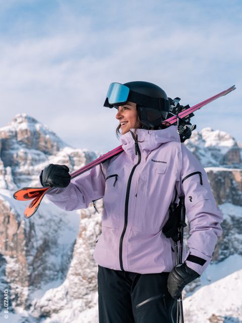 Une skieuse en veste lavande et casque sourit en portant ses skis sur l'épaule ; des montagnes enneigées sont visibles en arrière-plan.