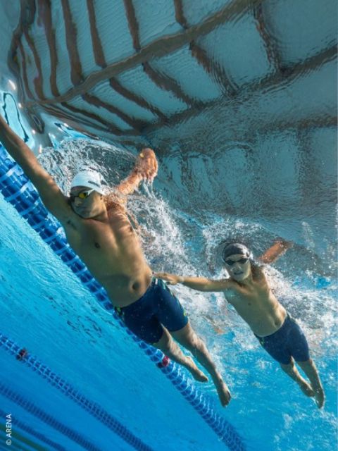 Deux nageurs avec des lunettes et des bonnets de bain font une course sous l'eau dans une piscine ; l'un tend la main vers l'autre près de la ligne de séparation des couloirs.