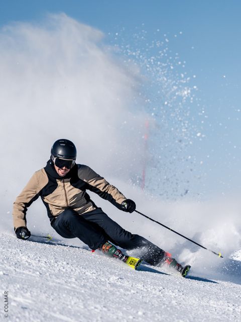 Un skieur avec un casque et une veste marron clair effectue des virages serrés sur une pente enneigée, soulevant de la poudreuse sous un ciel bleu éclatant.