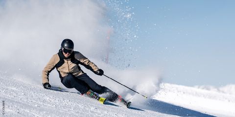 Un skieur vêtu d'une veste beige et d'un casque noir effectue des virages serrés sur une pente enneigée, soulevant un nuage de neige contre le ciel bleu éclatant.