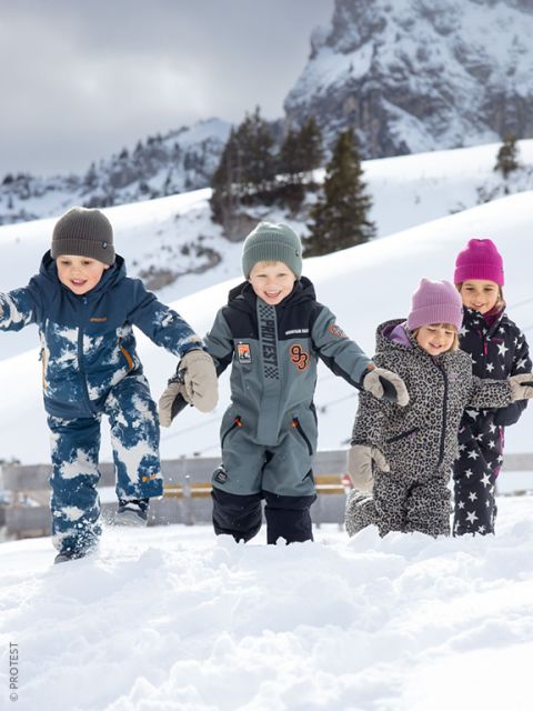 Quatre enfants en vêtements d'hiver jouent et sourient dans la neige, avec des montagnes et des arbres enneigés en arrière-plan.