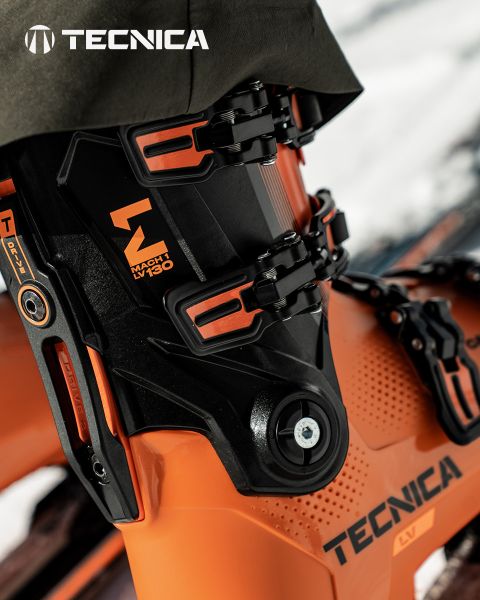 Un skieur en tenue rose vif et noire et casque blanc dévale une pente enneigée, se frayant un chemin dans la neige. En arrière-plan, des sommets majestueux enneigés s'élèvent sous un ciel légèrement nuageux. Dans le coin supérieur gauche, on peut lire le mot « Tecnica ».
