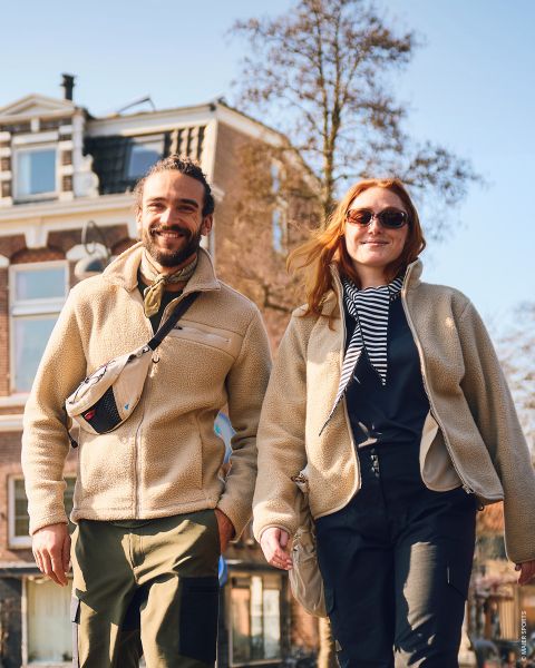 Un homme et une femme souriants se promènent en plein air par une journée ensoleillée, vêtus de vestes et de bottes décontractées, se tenant la main, avec un grand bâtiment en briques en arrière-plan.