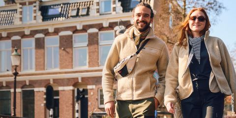 Un homme et une femme souriants se tiennent la main et traversent un pont par une journée ensoleillée, avec des bâtiments historiques en arrière-plan.