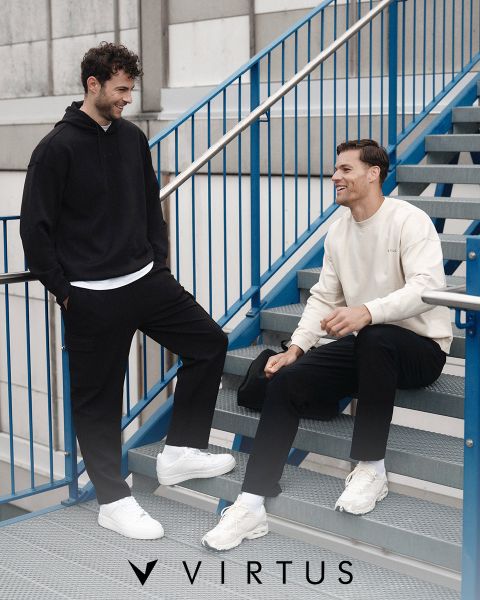 Deux hommes en tenues de sport Virtus, l'un portant un sweat à capuche gris clair avec un sac de sport noir, l'autre une veste noire, sourient et se promènent ensemble dans un hall moderne. Le logo Virtus est visible au-dessus d'eux.