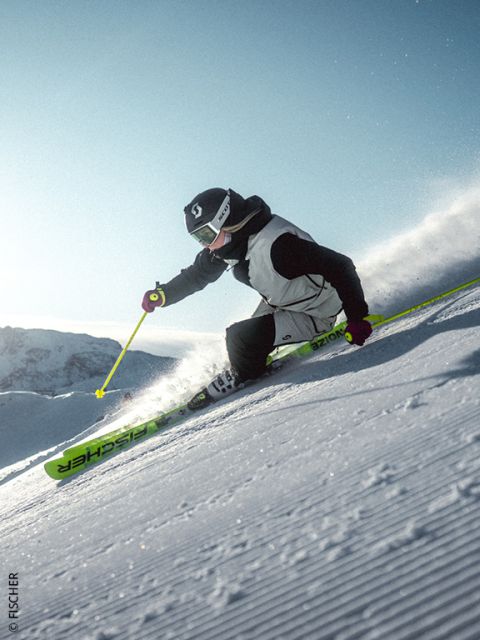 Un skieur en veste bleue et avec un casque dévale une pente de montagne enneigée au coucher du soleil, soulevant de la poudreuse, avec des sommets montagneux escarpés et ensoleillés en arrière-plan.