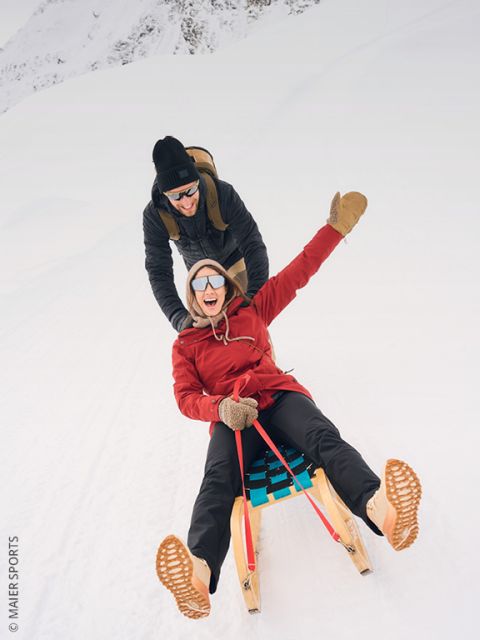 Deux personnes en vêtements d'hiver tirent un traîneau en bois sur une colline enneigée, avec des arbres et un sommet de montagne ensoleillé en arrière-plan. On aperçoit d'autres personnes au loin.