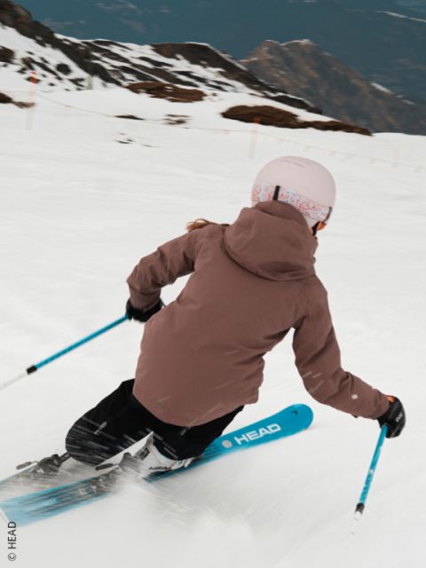 Un skieur en veste beige, pantalon bleu et casque blanc dévale une piste enneigée, effectue un virage serré et soulève de la neige. Des montagnes sont visibles en arrière-plan.